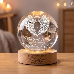Pode incluir: Uma lâmpada de esfera de vidro transparente com um design gravado de asas de anjo e uma auréola, com o texto "On Angel Wings you were taken away, but in our heart you will always stay." A base de madeira está gravada com "IN LOVING MEMORY OF Evelyn 1948-2026."