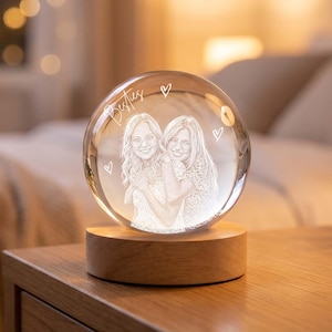 May include: A clear glass sphere featuring an internal laser-etched portrait of two people embracing. The word Besties and three small heart outlines are etched above the figures. The sphere rests on a circular, light-colored wooden base.