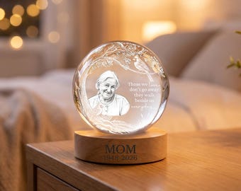 May include: A clear glass sphere featuring an etched portrait of an older person, a cardinal bird, and tree branches. The text reads: Those we love don't go away they walk beside us everyday. The sphere sits on a wooden base engraved with MOM 1948-2026.