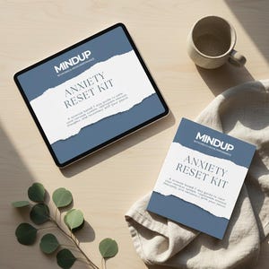 May include: A tablet and a booklet with the text "MINDUP ANXIETY RESET KIT" are displayed on a wooden surface. The tablet is black with a white and blue design. A small ceramic mug and a sprig of eucalyptus are also present.