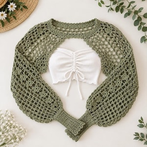 Puede incluir: Un bolero de ganchillo de manga larga en color verde salvia con un delicado patrón floral y puños acanalados, superpuesto sobre un top tubo blanco acanalado con detalle fruncido central y cordones ajustables en el frente.