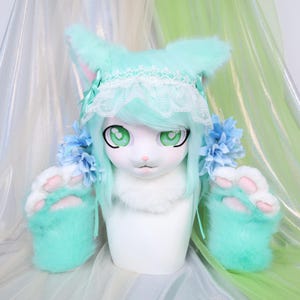 May include: A mint green and white furry cat head and paw costume set. The head has large green eyes, mint green hair, and a white face. The paws are mint green with pink paw pads. The costume is displayed on a white mannequin head.