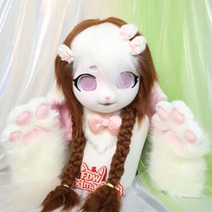May include: A white and brown bunny fursuit head with pink accents, large eyes, and long brown braids. The fursuit includes matching white paw gloves with pink paw pads. A pink bow tie and small pink bows adorn the head.