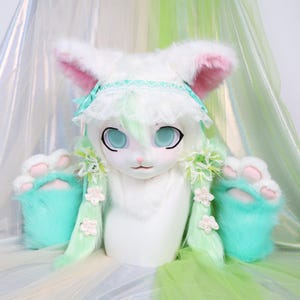 May include: A white furry costume head with large blue eyes, pink-lined ears, and a mint green and white wig. The costume includes matching mint green paw-shaped hand covers. The headpiece has a delicate lace and ribbon headband.