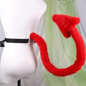 May include: A bright red faux fur devil tail attached to a white plastic base with an adjustable black nylon strap and a plastic side-release buckle. The tail features a curved shape with a pointed, arrow-like tip covered in long, vibrant red synthetic fur.