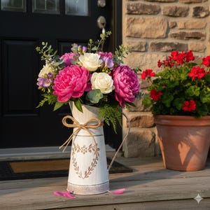May include: A white decorative jug with a heart design, filled with a vibrant floral arrangement. The bouquet includes pink peonies, white roses, and purple freesia. A brown bow adorns the jug, which sits on a wooden porch near a red geranium plant.