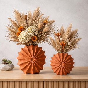 Może przedstawiać: Dwa wazony w kolorze terakoty z motywem promieni słonecznych zawierają suche kompozycje kwiatowe. Większy wazon zawiera mieszankę beżowej trawy pampasowej, pomarańczowych kwiatów i białych hortensji. Mniejszy wazon ma podobną kompozycję.