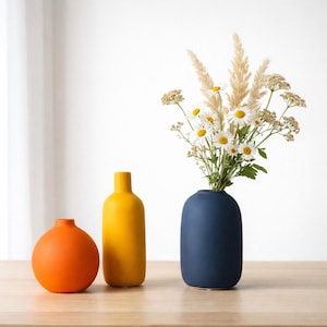 Könnte beinhalten: Drei Keramikvasen in einer Reihe. Eine orangefarbene Kugelvase, eine gelbe flaschenförmige Vase und eine marineblaue Vase mit weißen Gänseblümchen und hohem Gras. Die Vasen stehen auf einer hellbraunen Holzoberfläche.