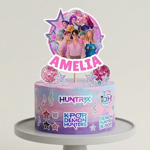 May include: A round cake with a pastel purple, blue, and pink ombre design. The cake is decorated with a K-Pop theme, featuring a group of cartoon characters, disco balls, stars, and the name "AMELIA". The cake also has the text "HUNTRX" and "K-POP DEMON HUNTERS".