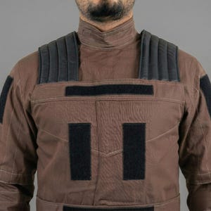 Op de afbeelding: Bruin tactisch vest met zwarte gewatteerde schouderaccenten en zwarte klittenband patches. Het vest heeft een hoge kraag en een gewatteerd ontwerp. De algehele esthetiek suggereert een militair of tactisch thema.