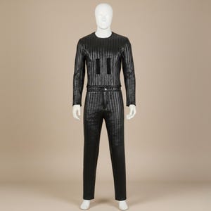 Puede incluir: Mono negro con rayas verticales sobre un maniquí. La parte superior de manga larga tiene dos acentos rectangulares negros. Los pantalones tienen un patrón de rayas verticales a juego y un cinturón. El maniquí es blanco y no tiene rasgos faciales.