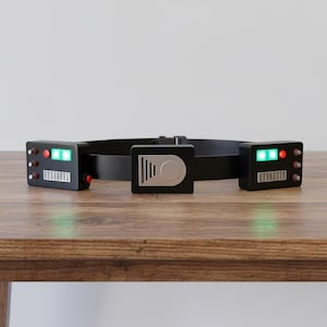 May include: A black belt with a square buckle and two rectangular side pieces. Each side piece has a series of buttons and lights, including green and red lights. The buckle has a silver design.