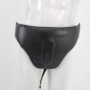 May include: Black leather garment with a curved waistband and a central, elongated panel. A black strap hangs from the bottom. The garment is displayed on a white mannequin.