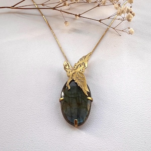 Puede incluir: Un collar de tono dorado con un colgante de piedra labradorita gris oscuro de forma ovalada. El colgante presenta una figura de ave de metal dorado posada sobre la piedra, asegurada con garras en una cadena fina a juego de tono dorado.