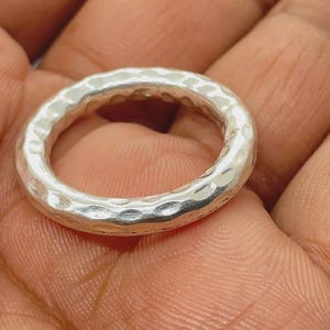May include: A silver-coloured ring with a hammered, textured surface. The band has a rounded, circular shape with a reflective metallic finish that catches the light, creating subtle highlights and shadows across its uneven, dimpled exterior.