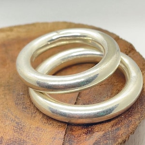 May include: Two silver-colored, thick, round rings are stacked on a wooden surface. The rings have a smooth, polished appearance, reflecting light. The rings are likely jewelry, possibly for fingers or other body parts.