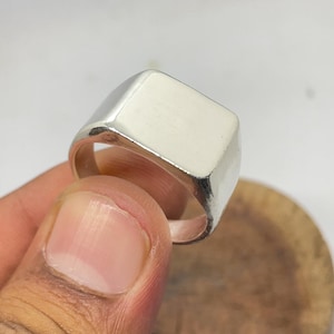 Puede incluir: Un anillo de sello plateado con una cara rectangular. El anillo está hecho de metal pulido y tiene una superficie lisa y reflectante. Una joya clásica.