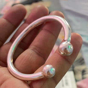May include: A silver bracelet with a curved design and two spherical ends. The open-ended bangle is shown in a close-up view. The bracelet is made of a shiny metal.