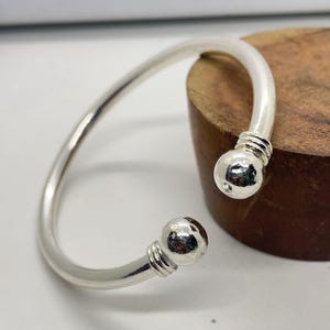 Puede incluir: Una pulsera de puño plateada con un diseño redondeado y abierto. La pulsera presenta una banda lisa y curvada que se ensancha en cada extremo en una bola esférica. Las bolas están acentuadas con un detalle texturizado y acanalado cerca de la banda.