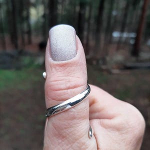 May include: A silver ring with a smooth, curved design is worn on a thumb with a light gray nail. The ring has a polished, reflective surface. The background is blurred, with a forest visible.