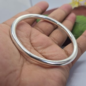 Könnte beinhalten: Ein silbernes Armreif-Armband mit glattem, abgerundetem Design. Das Armband ist aus massivem, poliertem Metall, das das Licht reflektiert. Der Armreif wird in einer Hand gehalten und zeigt seinen schlichten, eleganten Stil. Ein klassisches Schmuckstück.