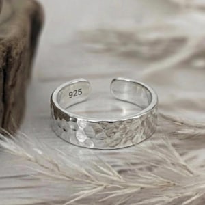 May include: A silver open-ended ring featuring a textured, hammered surface finish. The interior of the band is clearly stamped with the number 925, indicating the sterling silver composition of this adjustable piece of jewelry.