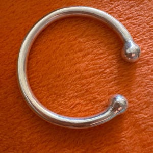Könnte beinhalten: Ein silberfarbenes, offenes Armreif-Armband. Das Armband hat ein abgerundetes, glattes Design mit einem kugelförmigen Ende auf jeder Seite. Das Armband wird vor einem orangefarbenen Hintergrund präsentiert.