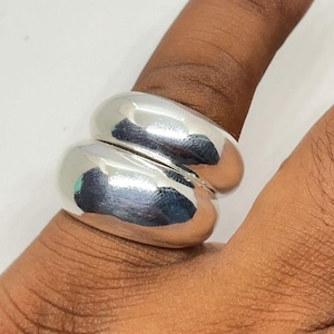 Puede incluir: Un anillo plateado brillante con un diseño único de doble banda. El anillo tiene una superficie lisa y pulida que refleja la luz. Se lleva en un dedo, mostrando su estilo elegante y moderno. Un accesorio llamativo.