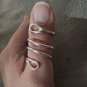 Può includere: Un anello color argento si avvolge attorno a un pollice. L'anello ha un design a spirale con due accenti a forma di goccia nella parte superiore e inferiore. L'anello è realizzato in metallo lucido e riflettente.