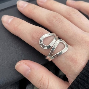 May include: A silver ring with a hammered texture. The ring features an open design with two overlapping teardrop shapes. The ring is worn on a finger, showcasing its unique, modern style. The metal has a bright, reflective quality.