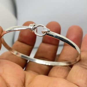 May include: A silver-colored bangle bracelet with a hook and eye closure. The bracelet is smooth and polished, with a simple, elegant design. The hook closure is a small, circular loop that connects to a curved bar.