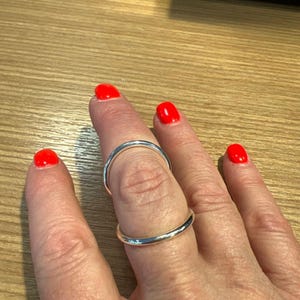 May include: A silver ring is worn on a finger with bright red nail polish. The ring is a simple band design, reflecting light. The background is a light-colored wooden surface.