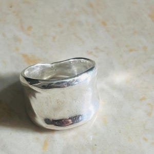 Peut inclure: Une bague argentée massive avec une surface lisse et irrégulière. La bague a une large bande et un bord supérieur légèrement inégal. Le métal reflète la lumière, créant des reflets et des ombres. Un bijou audacieux.