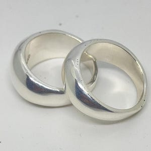 Könnte beinhalten: Zwei verbundene, breite Silberringe. Die Ringe sind glatt und glänzend und haben eine abgerundete Form. Die Innenseite der Ringe ist heller silberfarben. Die Ringe werden auf einer weißen Oberfläche präsentiert. Die Ringe scheinen aus Metall zu sein.