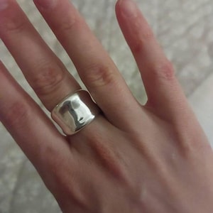 Può includere: Un anello largo in argento con una superficie liscia e riflettente. L'anello è indossato su un dito, mettendo in risalto il suo design audace e minimalista. La forma dell'anello è leggermente irregolare, aggiungendo un tocco unico al gioiello.