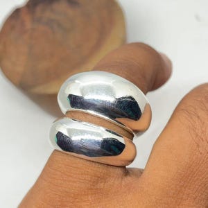 May include: A polished silver ring featuring a stacked, rounded design. The ring is worn on a finger, reflecting the light. The smooth surface of the ring contrasts with the skin's texture. The ring is a piece of jewellery.