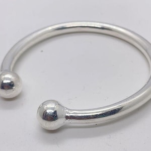 May include: A silver-colored open cuff bracelet with a rounded design. The bracelet features a smooth, polished finish and has a ball-shaped end on each side. The jewelry is displayed on a white surface.