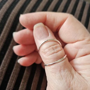 Può includere: Un anello d'argento indossato su un pollice. L'anello ha un design semplice e minimalista, con una sottile fascia che si curva attorno al dito. L'anello è lucido e riflette la luce. Lo sfondo è un tessuto a righe.
