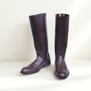 May include: A pair of dark brown knee-high boots with a smooth finish, rounded toes, and a low heel. The boots feature a tall, structured shaft that reaches the knee, with a small gold-toned snap button detail visible at the top inner edge.
