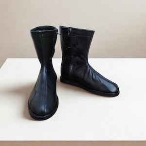 May include: A pair of black leather boots. One boot features a zip, the other has three buckles. The boots are mid-calf length and have a rounded toe. The boots are displayed on a white surface.