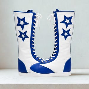 May include: A pair of white and blue knee-high boots. The boots have a white upper with blue star accents and a blue toe and heel. White laces run up the front of the boots.
