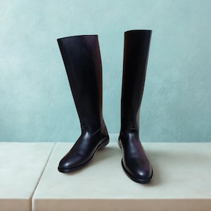May include: A pair of tall, black leather riding boots. The boots are knee-high with a sleek, minimalist design. The boots are displayed on a white surface against a light blue background.