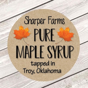 May include: A round label with the text "Sharper Farms PURE MAPLE SYRUP" in black. Two orange maple leaves flank the word "PURE". The text "tapped in Troy, Oklahoma" is at the bottom. The label is on a light brown background.