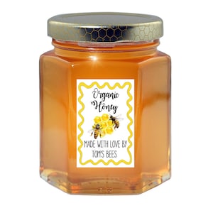 May include: A glass jar of organic honey with a yellow and white label that reads "Organic Honey" and "Made with love by Tom's Bees". The label has a honeycomb design with two bees.