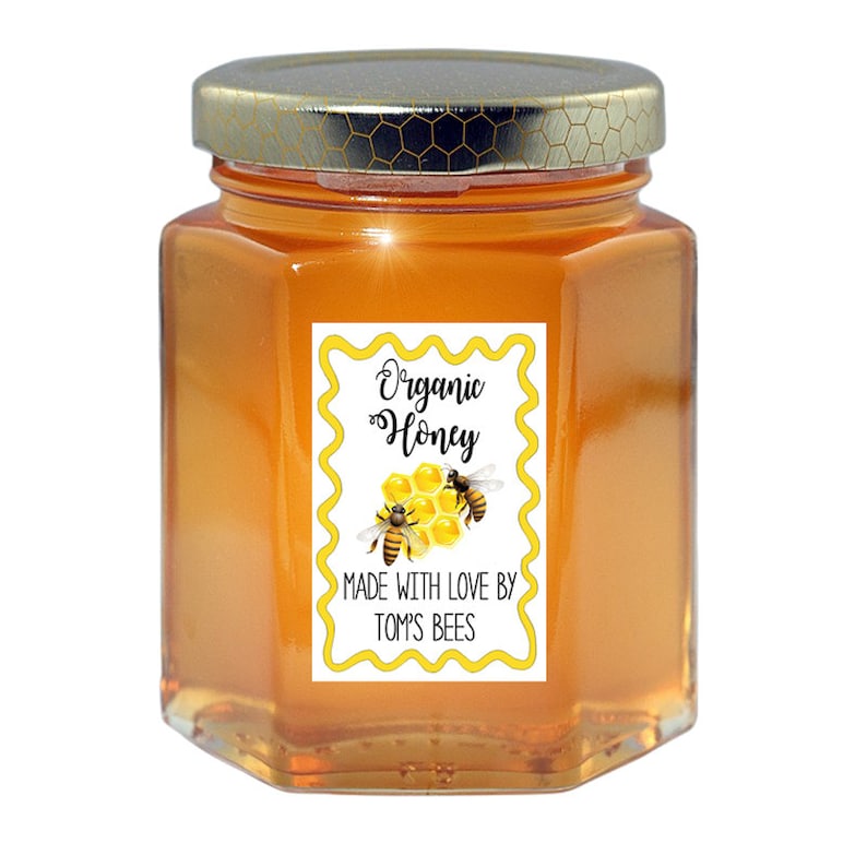 May include: A glass jar of organic honey with a yellow and white label that reads "Organic Honey" and "Made with love by Tom's Bees". The label has a honeycomb design with two bees.