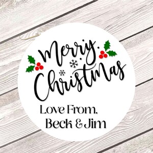 May include: A white circular sticker with the words "Merry Christmas" in black script, adorned with green holly leaves and red berries. The text "Love From, Beck & Jim" is below the main text. The sticker is set against a light wood grain background.