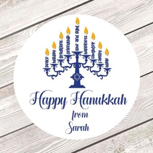 May include: A white circle sticker with a blue menorah and the words "Happy Hanukkah from Sarah". The menorah has eight candles with yellow flames and the words "peace", "holiday", "traditions", "friendship", "love and light", "happiness", "laughter", "family", and "home" around it.