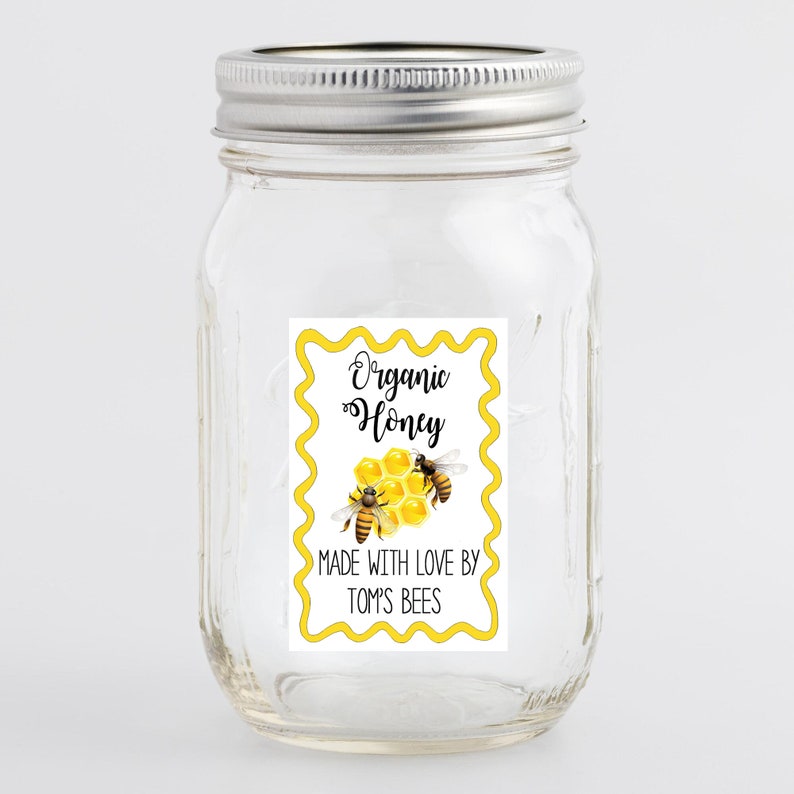 May include: A white label with yellow border and wavy edges. The label says "Organic Honey" in black cursive font. Below the text is a drawing of a honeycomb with two bees. The label also says "Made with love by Tom's Bees".
