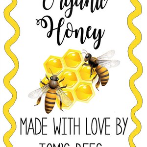 May include: A yellow and white label with a wavy border. The label says "Organic Honey" in black cursive text. There are two cartoon bees on the label, one on each side of a honeycomb. The text "Made with Love By Tom's Bees" is below the honeycomb.