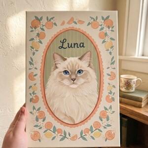 May include: A rectangular canvas print featuring a portrait of a fluffy cream-colored cat with blue eyes inside an oval frame. The name Luna is written above the cat. The border is decorated with illustrations of orange slices and white flowers.